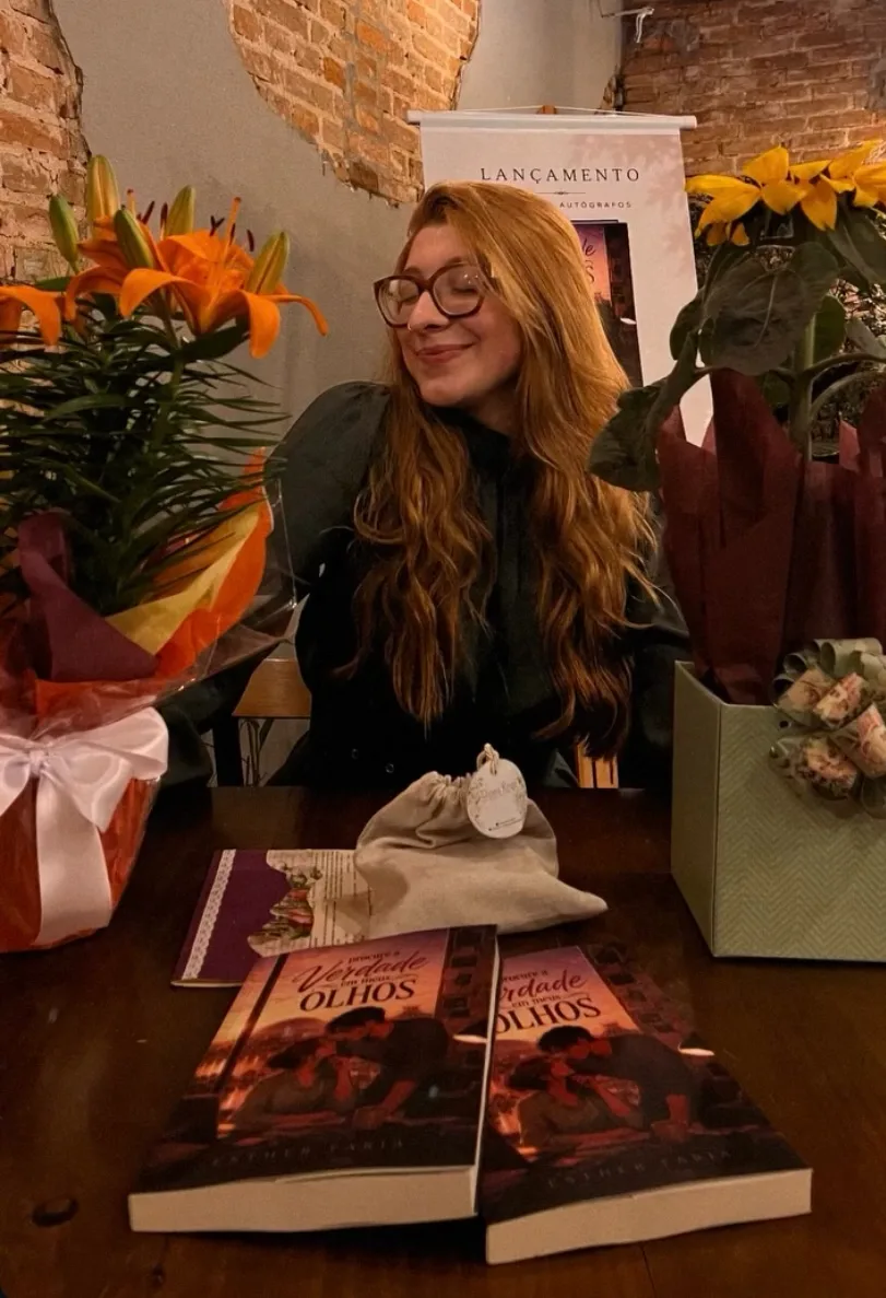 Esther Faria sentada à mesa de autógrafos, cercada de flores e exemplares